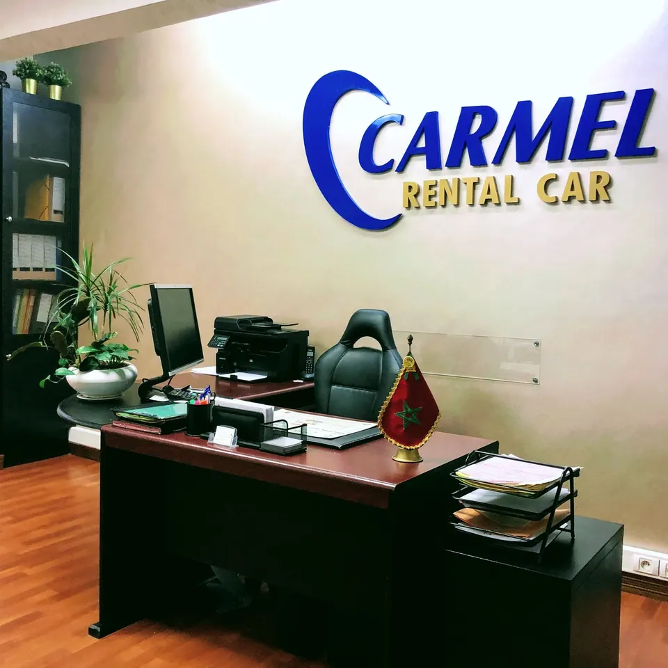 Bureau de l’agence avec le logo CARMEL RENTAL CAR en relief bleu et orange au mur